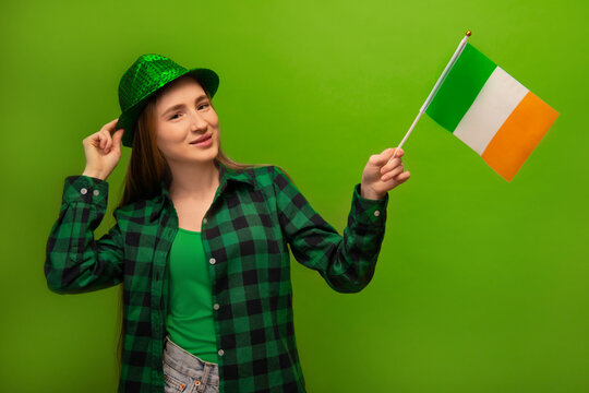Happy cheerful smiling irish woman in green checkered plaid shirt and sparkly shiny party hat holding small Ireland national flag isolated on colorful green background.

St Patricks Day concept.