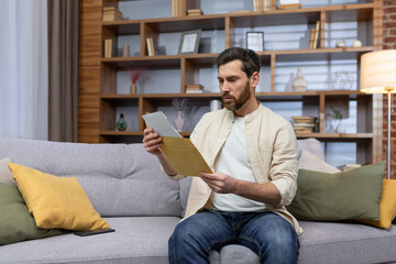 Upset young man sitting on sofa at home, received a letter, reads bad news. Divorce documents,...