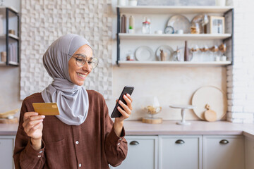 Young smiling Arab woman in hijab using phone and credit card at home in kitchen. Do online...