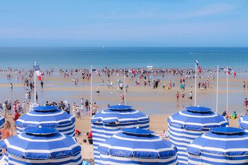 A normandy beach with rows of white and blue parasols
at low tide and a crowd of bathers