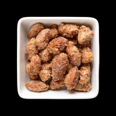 Candied almonds in a white square bowl. Homemade, in a special way cooked almonds, whole nuts coated in crunchy sugar. Sold at Christmas markets. Close-up, from above, isolated over black, food photo.