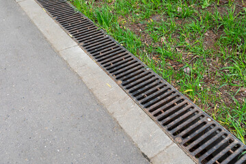 A rusted metal sewer grate along a paved walkway. The edge of the pavement