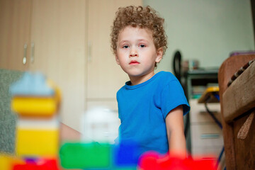 Little curly boy plays with toys. The child builds a house from a colored constructor.