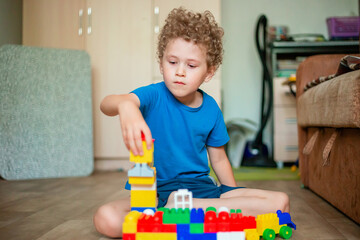 Little curly boy plays with toys. The child builds a house from a colored constructor.