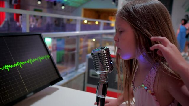 Playful girl sings and screams into microphone emotionally. Junior schoolgirl looks at moving sound wave on monitor on blurred background closeup