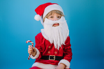 Fototapeta na wymiar Funny boy in Santa Claus hat and sweet caramel cane on blue studio background