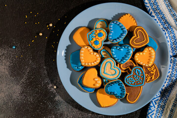 Cookies in shape of hearts decorated by yellow and blue icing and sprinkles and towel embroidered with Ukrainian ornament on dark background