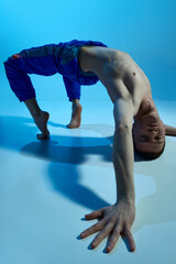 Fototapeta premium Contemporary dance style. Young artistic man dancing contemp, experimental dance over blue studio background. Flexible artist. Concept of art, body aesthetics, motion, action, inspiration.