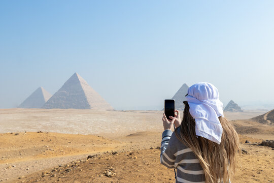 Unrecognizable young tourist woman from the back with white turban on head taking a picture of the Pyramids of Caire, Egypt