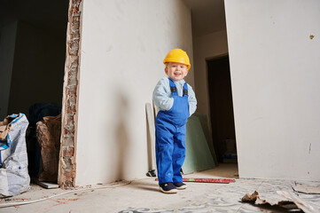 Full length of cheerful little boy in safety construction helmet standing by the wall in apartment under renovation. Joyful kid in work overalls looking at camera and smiling. Home renovation concept.