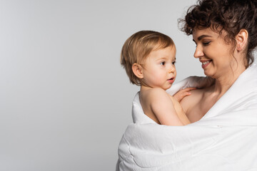 happy mother looking at toddler baby girl covered in duvet isolated on grey.