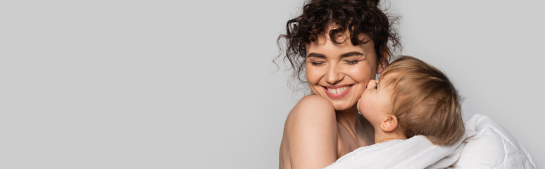 toddler baby girl kissing cheek of cheerful mother covered in white duvet isolated on grey, banner.