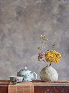 White ceramic teapot and cup of fresh hot beverage placed on wooden table near vase with dried flowers against gray wall