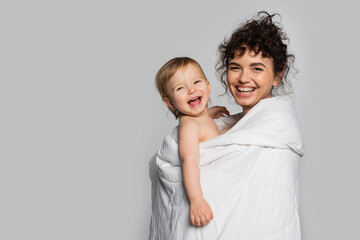 happy mother and positive toddler daughter covered in duvet isolated on grey.