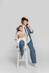 happy businesswoman in blue suit talking on smartphone and sitting with toddler daughter on high chair on grey.