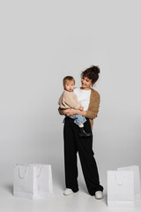 full length of cheerful mother in casual clothes holding in arms toddler baby near shopping bags on grey.