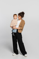 full length of stylish mother in casual clothes holding in arms baby girl in sweater on grey.