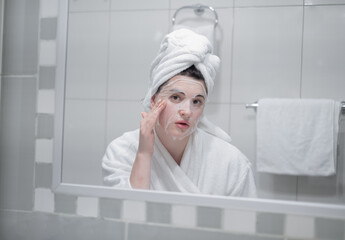 Young caucasian woman in a white bathrobe and with a towel on her head removes a moisturizing sheet mask from her face, beauty treatments in the bathroom