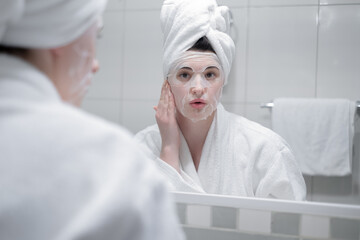 Young caucasian woman in a white bathrobe and with a towel on her head removes a moisturizing sheet mask from her face, beauty treatments in the bathroom