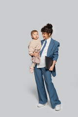 full length of cheerful businesswoman in blue suit holding in arms toddler daughter and laptop on grey.