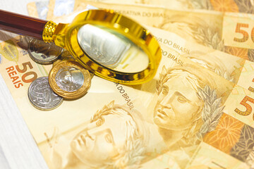 Notas do Real Brasileiro sobre uma mesa com uma lupa dourada na composição. Economia brasileira e...