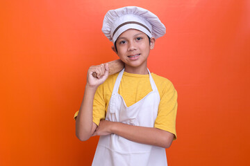 Smiling little boy in chef's toque and apron holding wooden rolling pin on shoulder over orange background.