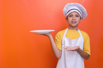 Smiling little boy in chef's toque and apron holding empty plate while doing presenting gesture on...