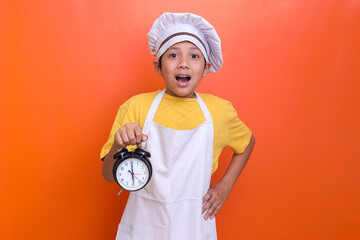 Cute boy wearing chef uniform gesturing shocked expression while holding alarm clock isolated on...