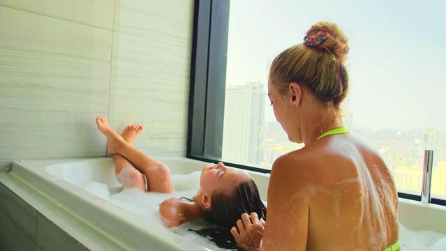 Young mother and little daughter take a bath in bathroom. Family of one parent with child. Concept growing up, childhood, motherhood, domestic life. Cute mom and kid take a bath with foam in bathtube