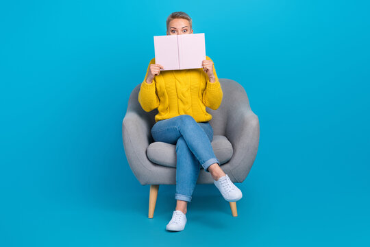 Full length photo of funky girl sit comfy armchair enjoy free time spending hide face silly reading literature isolated on blue color background
