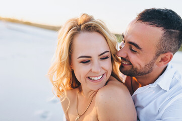 A tanned young caucasian bearded guy kisses a beautiful fun smiling blonde woman against the backdrop of the setting sun. Desert, sandy beach, rest and relaxation. Honeymoon concept.