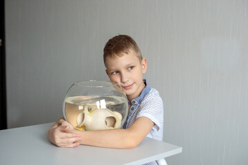 A cute boy sits near a transparent aquarium in the room and hugs an aquarium with fish