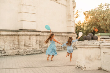 Obraz na płótnie Canvas Daughter mother run holding hands. In blue dresses with flowing long hair, they hold balloons in their hands against the backdrop of a sunset and a white building.