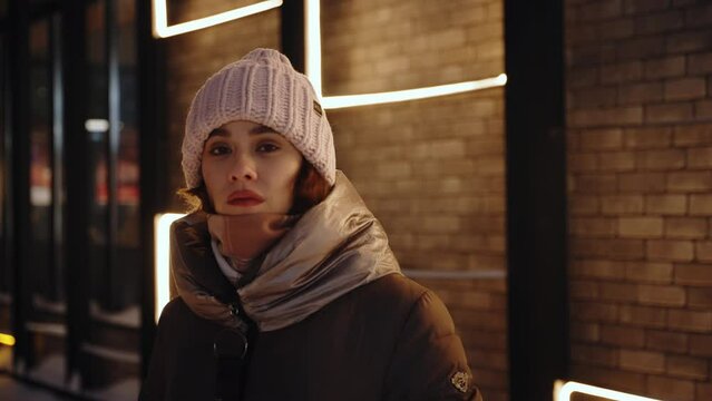 A sad girl stands in the cold and looks into the camera. Beautiful young woman in a winter hat and coat walking in the city at night.