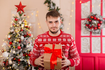 Obraz na płótnie Canvas A young man in a red sweater in Scandinavian style holds a box with a gift in his hands. The guy on the background of the Christmas tree porch. Surprise