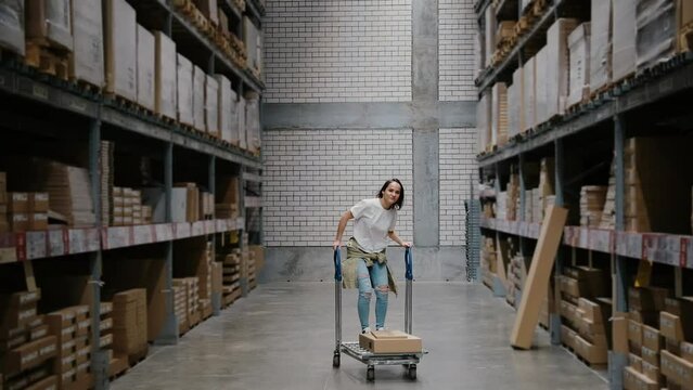 Girl rides shopping cart in furniture store warehouse between the rows of shelves with cardboard boxes