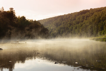 Silent beautiful morning summer landscape - mist on river with warm golden fluffy haze on water with reflection, lush green forest on slopes in pink sunrise sunbeams. Majestic wild nature for travel.