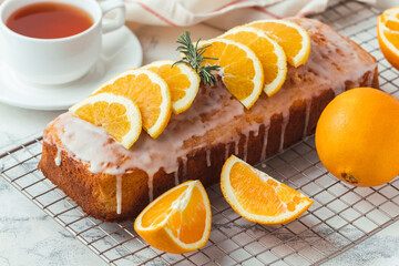 Obraz na płótnie Canvas Loaf of orange bread covered with a confectionery glaze with lemon juice and decorated with orange slices. Chiffon cake on a pastry grill next to cup of tea. White background