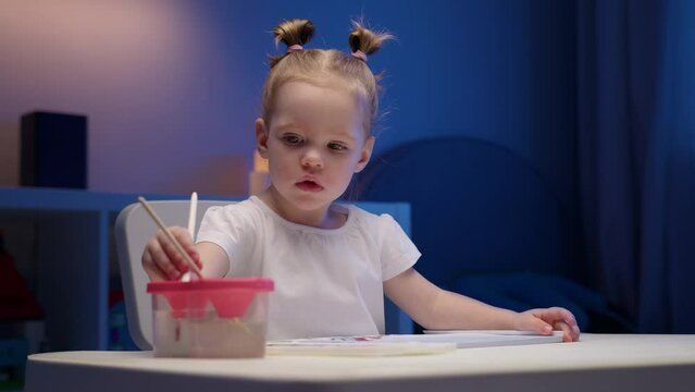 Cute two-year-old blonde girl paints multicolored paints on white paper sitting at home at table in evening. Child paints with watercolor paints sitting at table in evening.