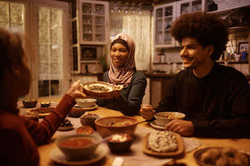 Happy Muslim woman passing food to her daughter during family dinner at dining table.