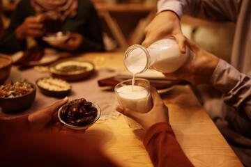 Close up of Muslim father pouring milk into daughter's glass during family meal at dining table.