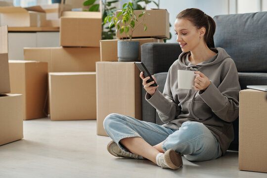 Young woman having video call on smartphone and drinking coffee during her rest after relocation