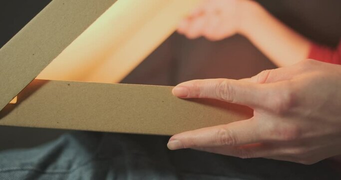 Women's hands open a cardboard pizza box. A bright yellow light comes out of it. Close-up, indoors, side view.