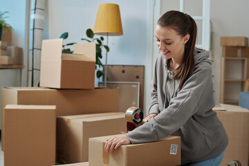 Smiling young woman packing cardboard boxes with adhesive tape to move to a new house