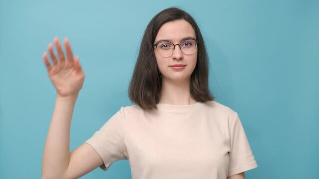 Portrait of a student girl with glasses and a beige T-shirt waving goodbye on a blue background, 4K. Bye-bye. See you soon. Nonverbal communication