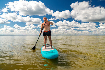 A man in sports shorts with a paddle stands on a SUP board in the lake against the background of white clouds on a clear blue sky.