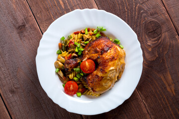 Pasta with vegetables and baked chicken leg with tomatoes sprinkled with green onions in a plate on the table.