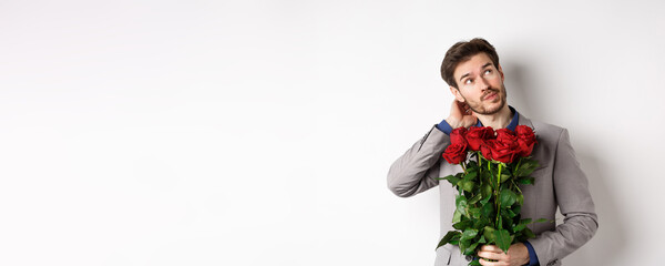 Thoughtful young man in suit going on valentines day date, holding bouquet of roses, thinking and...