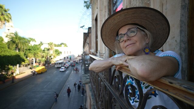 Closeup Wide Angle View Of Pretty Mature Senior Woman Wearing Hat, Ethnic Blouse Leaning On A Railing At A Cafe Restaurant Overlooking The City In Europe Or Latin America, Happy To Be Traveling.