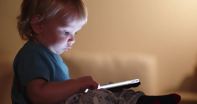 Little Child looking on mobile phone screen. Small child pressing with finger on phone touch screen. Light from the mobile phone screen illuminates the face of a small child. Cell phone and child